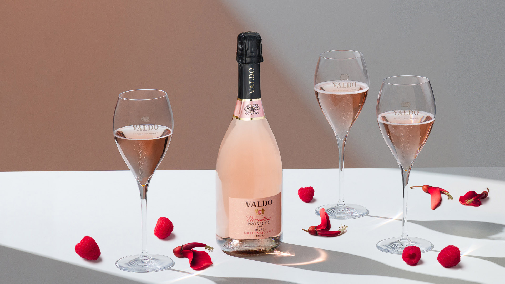 Bottiglia di Valdo Elevantum Prosecco DOC Rosé affiancata da tre calici colmi di spumante rosé, decorati con lamponi freschi e petali di rosa su un tavolo bianco ambientazione per san Valentino